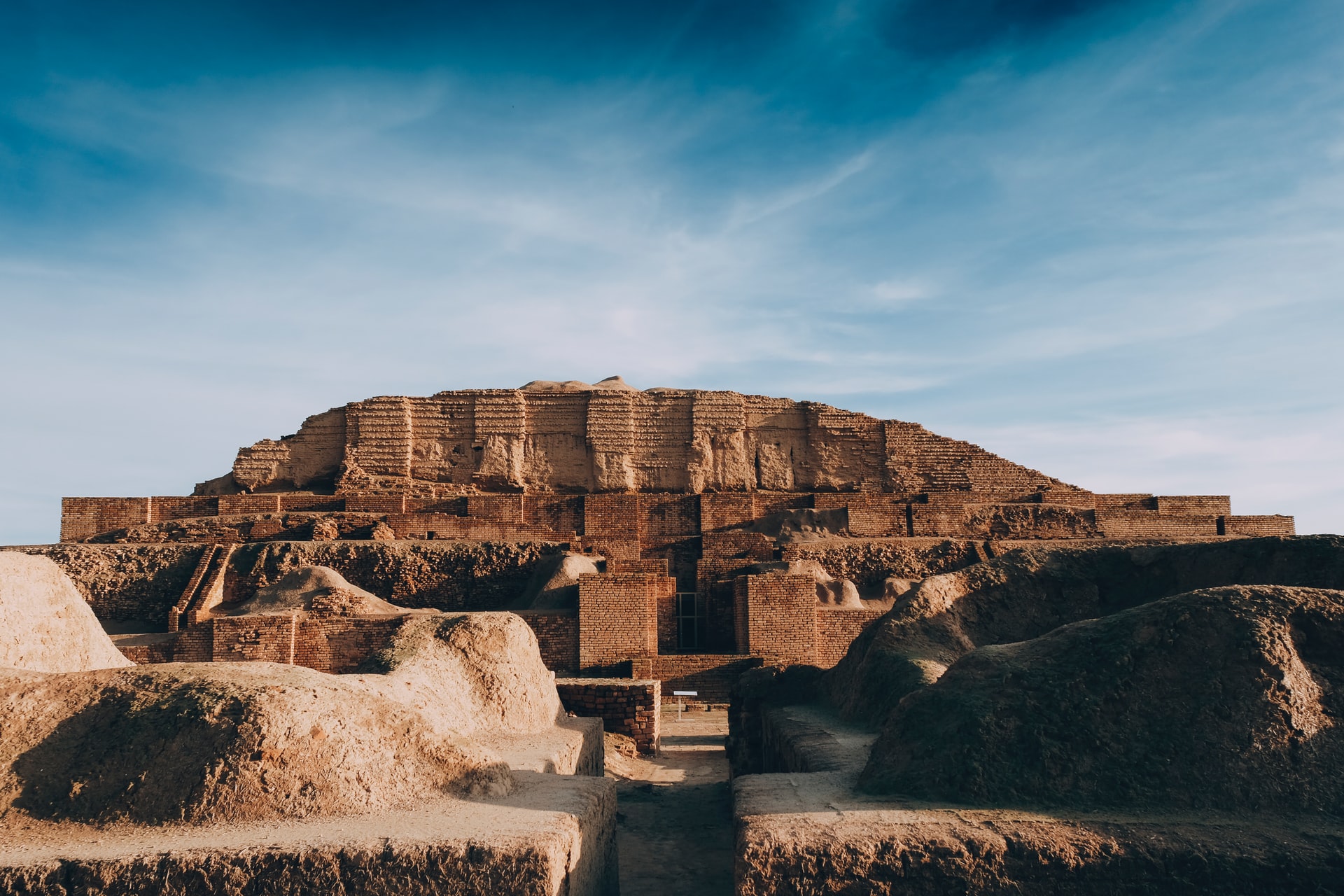 Ancient ziggurat pyramid against a blue sky, constructed from weathered, reddish-brown stone.